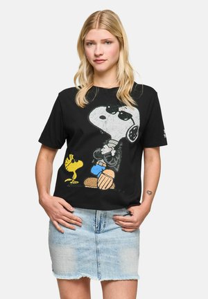 Woman with long blonde hair wears a black Snoopy and Woodstock T-shirt and a light blue denim skirt, standing with hands on hips.