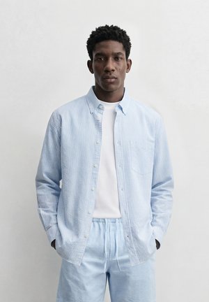 Rövid, göndör hajú férfi világoskék csíkos gombos ingben, fehér pólóval alatta, és hozzáillő világoskék csíkos nadrágban, kezét zsebében tartva.