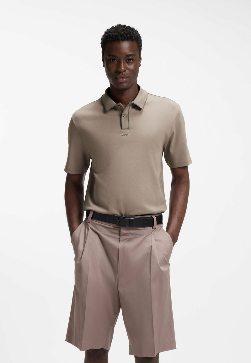 Homme portant un polo beige à manches courtes, rentré dans un short plissé marron clair avec ceinture noire, debout les mains dans les poches sur fond blanc.