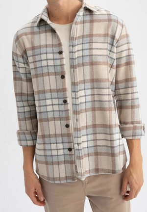 Homme portant une chemise à carreaux beige, bleu clair et marron boutonnée sur un t-shirt blanc, manches retroussées, et pantalon beige.