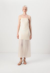 Robe sans manches de couleur crème avec une texture pailletée, corsage ajusté et jupe transparente effleurant le sol. Associée à des sandales beiges à lanières.