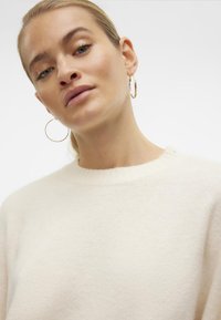 Cremefarbener Pullover mit einer weichen, flauschigen Textur und geripptem Rundhalsausschnitt, ergänzt durch große Creolen.