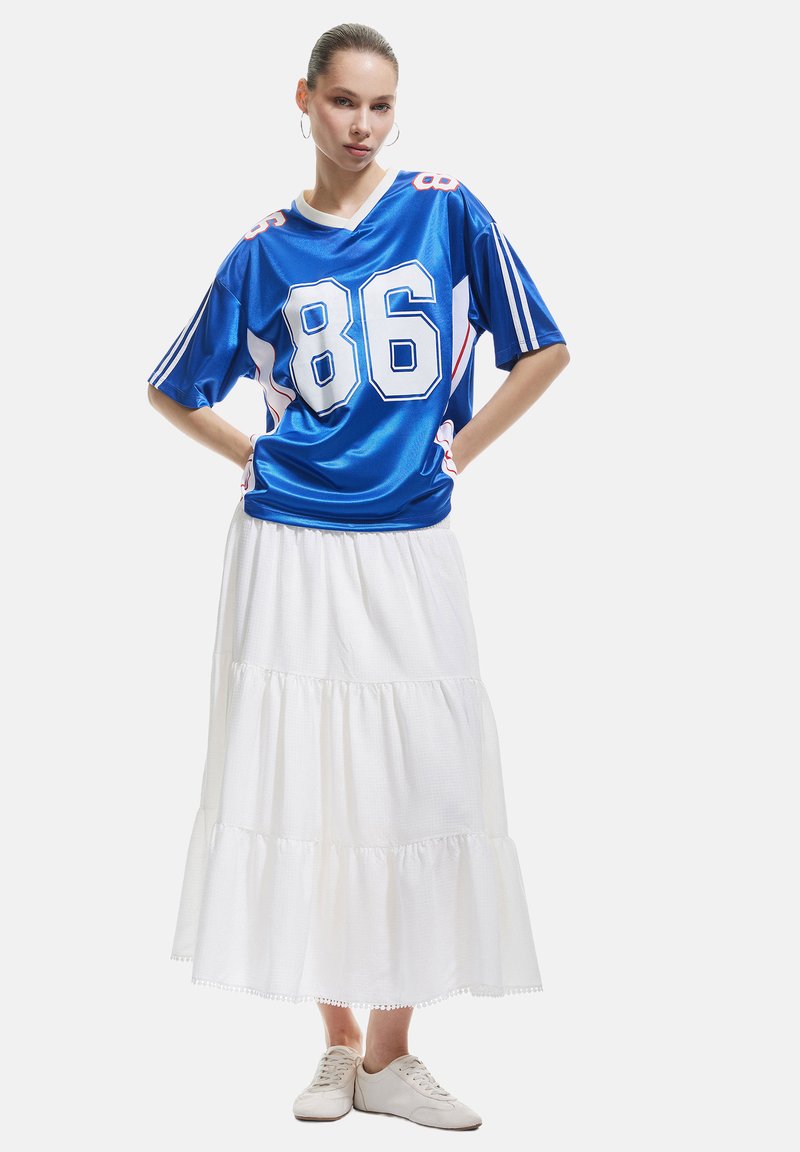 Blå satinjersey med hvide og røde detaljer, med nummer "86" på fronten, parret med en hvid lagdelt nederdel og lyse sneakers.