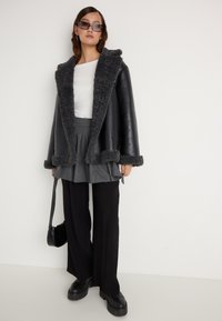 Jachetă din piele ecologică neagră cu detalii din blană ecologică gri, asortată cu o fustă plisată gri și pantaloni largi negri. Accesoriile includ ochelari de soare închiși la culoare și o geantă mică.