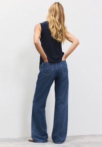 Kvinde med langt blondt hår står med front mod væggen, hænder i baglommerne på blå vidde-bukser, iført navy ærmeløs top og sandaler.