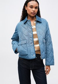 Kvinde iført en blå quilted jakke over en beige og hvid stribet sweater og mørke jeans, stående foran en lys ensfarvet baggrund.