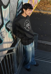 Schwarzer Kapuzenpullover mit rundem Celtics-Logo und weißem Text, kombiniert mit blauen, ausgewaschenen Jeans und schwarzen Schuhen, getragen vor einem bunten Graffiti-Hintergrund.