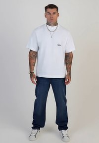 Camiseta blanca de manga corta con logo bordado, combinada con vaqueros azul oscuro. La modelo lleva zapatillas blancas y accesorios plateados.