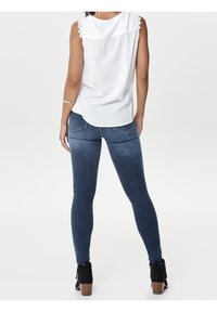 Jeans in denim blu, attillati con vita media, dotati di tasche posteriori e un leggero sbiadimento. Abbinati a una maglietta bianca senza maniche.