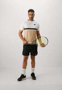 Tenisový hráč v bielo-beige tričku so zónovým vzorom a námorníckym pruhom, čiernych šortkách, bielych ponožkách a čiernych teniskách Nike, drží raketu.