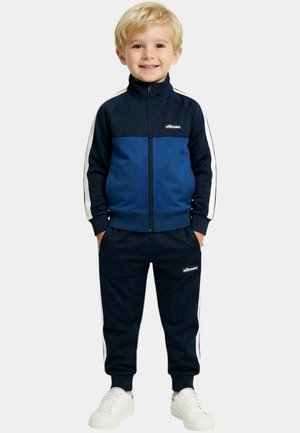 Jeune garçon souriant, vêtu d'un survêtement zippé bleu marine et bleu avec des bandes blanches et des baskets blanches, debout les mains dans les poches.