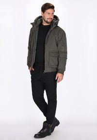 Hættejakke i olivengrøn farve med polstring, foran lynlås, to store sidelommer og ribkantsmanchetter, båret over en sort skjorte og sorte bukser.