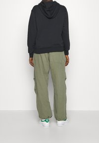 Svart huvtröja med elastiska ärmslut i kombination med löst sittande olivgröna cargobyxor. Vita sneakers med gröna detaljer.