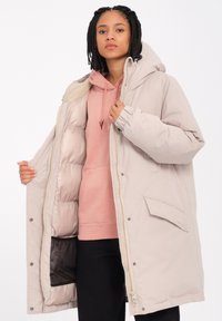 Beige parka jakke med hætte, lynlås lukning, frontlomme og quiltet foret. Båret over en lyserød hættetrøje med en blød tekstur.