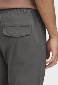 Grijze broek met elastische taille en een achterzak, met een gladde textuur en een subtiele glans, weergegeven vanaf de achterkant met een hand die ernaast rust.