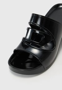 Sandal de slide preto e brilhante com uma larga fita apresentando um design sulcado, biqueira aberta e sola de plataforma robusta. Textura suave.