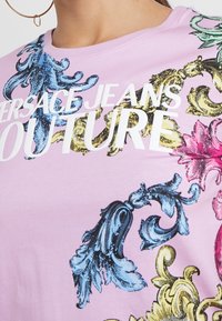 Růžové tričko s propracovanými modrými, žlutými a růžovými barokními vzory. Tučný bílý text „VERSACE JEANS COUTURE.“ Měkký bavlněný materiál.