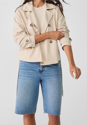 Persoon draagt een beige double-breasted jas met opgerolde mouwen en lichtblauwe denim knielange shorts.