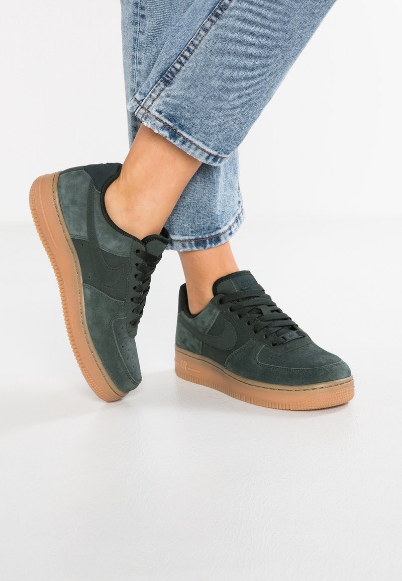 Personne portant des baskets en daim vert foncé à semelles en gomme et lacets noirs, associées à un jean bleu clair retroussé devant un fond blanc.