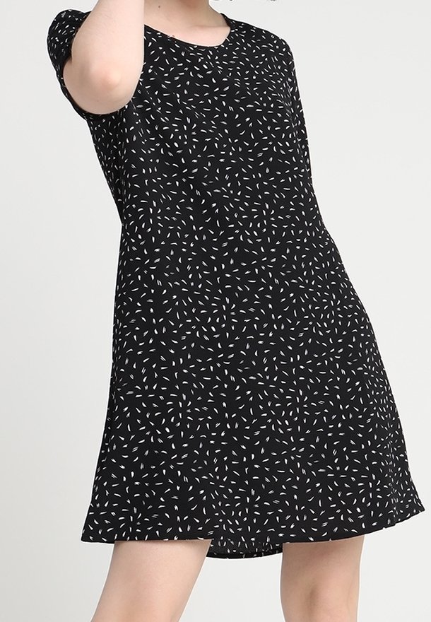 Femme portant une robe noire à manches courtes avec des motifs abstraits blancs en forme de traits, debout devant un fond clair uni.