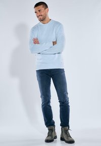 Hellblauer gestrickter Pullover mit geripptem Saum, kombiniert mit blauen Jeans und grünen Wanderschuhen. Das Modell steht mit verschränkten Armen.
