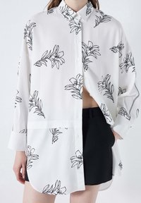 Camicia bianca con bottoni, decorata con contorni floreali neri, maniche lunghe e orlo curvo. Abbinata a pantaloni corti neri per creare contrasto.