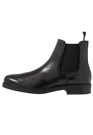 Botín Chelsea de piel negra con panel lateral elástico y lengüeta para tirar, tacón bajo, diseño elegante y logo de Calvin Klein grabado cerca del tobillo.