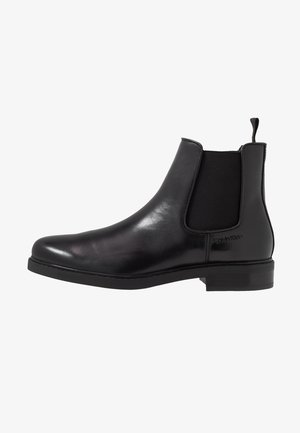 Bottine Chelsea en cuir noir avec panneau latéral élastique et languette, talon bas, design épuré et logo Calvin Klein embossé près de la cheville.