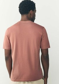 Camiseta de manga corta y cuello redondo en color rosa empolvado. Hecha de una tela suave, presenta una textura lisa y un corte relajado. Se muestra la vista trasera.