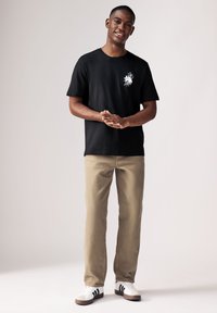 Schwarzes T-Shirt mit grafischem weißen Blumenmuster auf der Brust, kombiniert mit beigen Hosen und weißen Sneakers mit schwarzen Streifen.