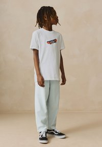 Weißes Baumwoll-T-Shirt mit einem bunten Skateboard-Motiv, kombiniert mit hellblauen, locker sitzenden Jeans und schwarzen Sneakern mit weißen Akzenten.