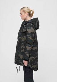 Kamuflāžas parka tumši zaļās un pelēkās krāsās, kurai ir kapuce, priekšējās kabatas un regulējams dzīslu šķipsnu apakšmalā.