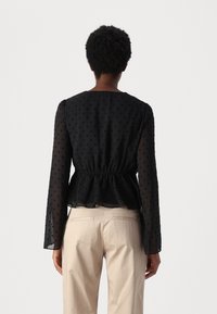 Donna con capelli corti e ricci che indossa una blouse nera a maniche lunghe con pois e pantaloni beige, vista da dietro su uno sfondo bianco.