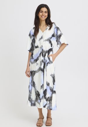 Lachende vrouw met donker haar, gekleed in een witte midi-jurk met zwarte en blauwe aquarelpatronen en bruine sandaaltjes met hak.