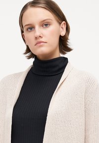 Lys beige strikket cardigan over en svart ribbestrikket høyhalset genser. Cardiganen har åpen front og teksturert stoff med løs passform.