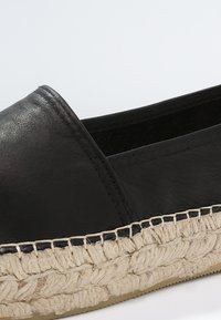 Chaussure espadrille en cuir noir sans lacets avec semelle en jute naturelle tressée et coutures détaillées en gros plan.