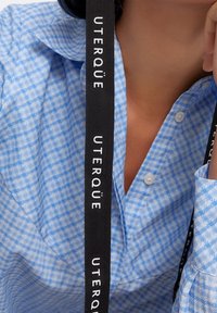 Camicia button-up azzurra chiara con motivo a diamante, dotata di un grande colletto e maniche lunghe. Cintura nera con la scritta "UTERQUË" in bianco.