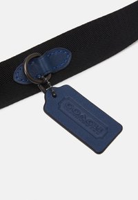 Ceinture en nylon noire avec une étiquette en cuir bleu portant un logo en relief, un anneau en métal rond et des détails en surpiqûre bleue.