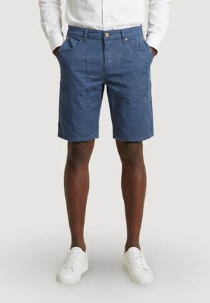 Hombre con pantalones cortos azules hasta la rodilla con bolsillos delanteros distintivos, camisa blanca abotonada y zapatillas blancas, de pie con las manos en los bolsillos.