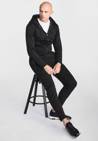 Czarna bluza z zamkiem błyskawicznym z białą podszewką, w połączeniu z pasującymi czarnymi dresami. Wyeksponowana na stołku, z delikatnymi niebieskimi akcentami.