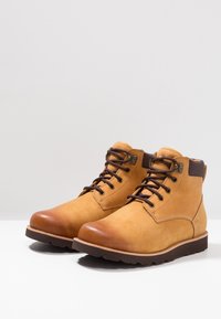 Botas de cuero marrón claro hasta el tobillo con cordones y suelas marrón oscuro, que cuentan con cuellos acolchados y ojales de metal para abrocharse sobre una superficie blanca.