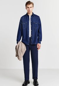 Camicia in denim blu con bottoni a pressione e due tasche sul petto, abbinata a pantaloni navy. Il modello tiene un maglione beige a coste. Scarpe nere.