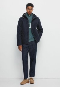 Giacca navy con cappuccio, chiusura con zip e tasche laterali, indossata sopra un maglione verde. Abbinata a pantaloni in denim scuri e scarpe beige.