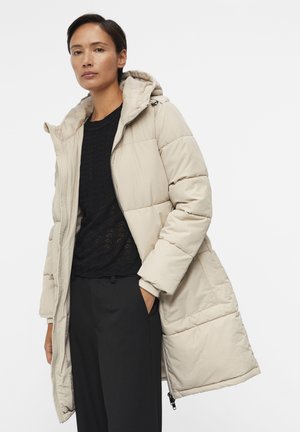 Manteau rembourré beige avec capuche, présentant des sections matelassées visibles. Porté sur un pull noir texturé et un pantalon foncé.