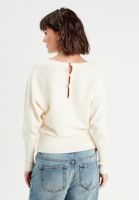 Cremefarvet sweater med vid halsudskæring og nøglehul i ryggen, med ribbede manchetter og kant. Kombination med blå denimjeans.