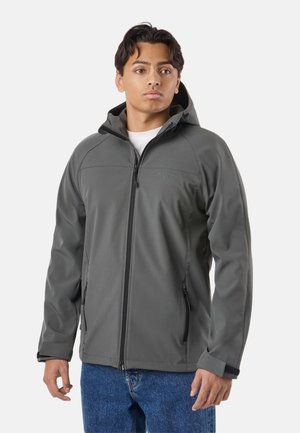 Jeune homme portant une veste softshell grise à capuche avec des poches zippées et un jean bleu, debout devant un fond blanc.