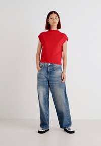 Rød kortermet topp med høy hals, kombinert med vide lyseblå jeans og sølvfargede platåsko, mot en ensfarget bakgrunn.