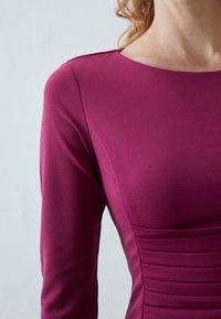 Úzké šaty v jednobarevné magentové tkanině, s dlouhými rukávy, kulatým výstřihem a vertikálním nařasením na trupu.