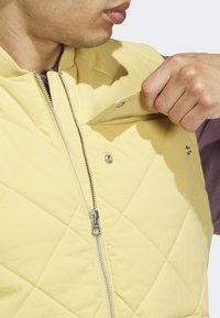 Muž rozopína žltú prešívanú vestu a dvíha západku pri hrudi, pod ňou má na sebe fialovú košeľu s dlhým rukávom, záber zblízka na hornú časť trupu.