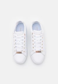 Pár bílých nízkých tenisek Tommy Hilfiger se zlatými očky a štítky "TOMMY" na tkaničkách, pohled shora na bílém pozadí.